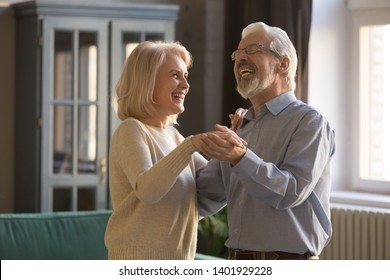 Laughing aged spouses holds hands standing in living room dancing at home, elderly family healthy active retirees celebrates anniversary chatting feels happy, forever love harmony in relations concept