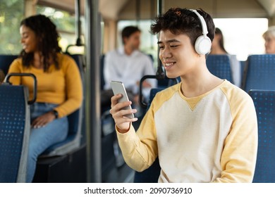 Laughing Asian Guy Using Mobile Phone Sitting On City Bus Seat, Happy Male Passenger In Wireless Headphones Listening To Music, Watching Video Or Movie, Making Online Call, Enjoying Travel Ride