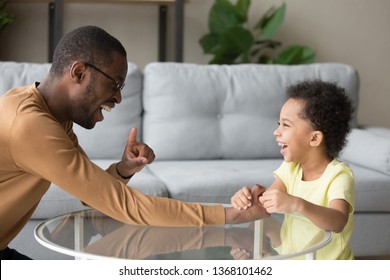 Laughing black father and biracial cheerful son having fun on weekend try to play explain arm wrestling game hold hands feels happy overjoyed. Concept of family play, activity at home, love upbringing