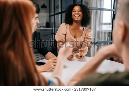 Laughing black female playing fun cards games for time with diverse friends, enjoying pastime activity sitting at table at home. Happy multiethnic men and women playing board game in living room.