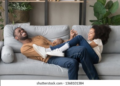 Laughing black funny father or tickle play with daughter preschool or schoolgirl kid girl lying together on sofa in living room at home, leisure lazy weekend activity game with child free time concept