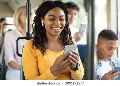 Laughing Black Lady Using Mobile Phone Sitting On City Bus Seat, Happy Passenger In Wireless Headphones Listening To Music, Watching Video Or Movie, Making Online Call, Enjoying Travel Ride