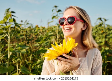 Laughing blond girl in sunglasses and white dress standing by one of sunflowers in front of camera against clear sky on sunny summer day - Powered by Shutterstock