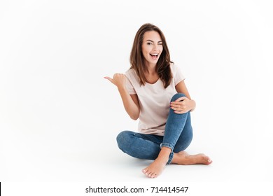Laughing brunette sitting on the floor, pointing away and winks at the camera over white background