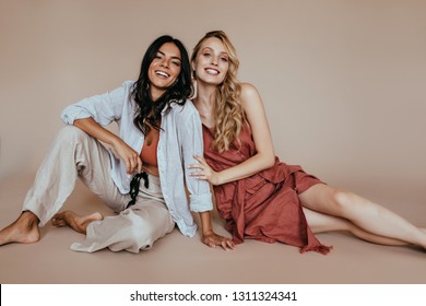 Laughing brunette woman in white pants sitting on the floor. Indoor photo of adorable female models posing together on light background.