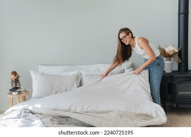 Laughing caucasian beautiful young woman makes bed wears jeans and white t-shirt and glasses. Pretty hispanic girl makes housekeeping. Mockup, people at home. Cheerful student makes order home. - Shutterstock ID 2240725195