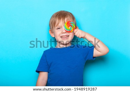 Laughing Caucasian child kid boy close one eye bright colorful lollipop candy. Close-up studio shot on blue background.