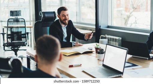 Laughing cheerful ambitious boss talking with colleagues while sitting at head of table during discussing business plan in meeting room - Shutterstock ID 1589712250