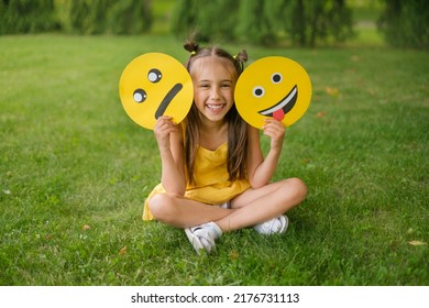 A Laughing, Cheerful Child Holds Two Emoticons In His Hands - A Sad, Upset One And A Smile Face Showing His Tongue