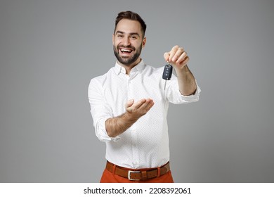 Ríe alegre y divertida joven empresario barbudo de 20 años con un puesto clásico de camisa blanca apuntando con las llaves del auto aislado en un retrato de estudio de fondo de color gris. Concepto de riqueza profesional