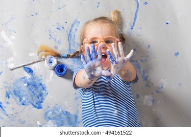 Laughing child with blue paint dots on face with colors and brushes lying on floor.