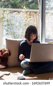 Laughing Child Sitting With Screen On Lap, Playing With Teddy Bear, Enjoying Homeschooling With Computer To Learn Online, Sunny Balcony Background