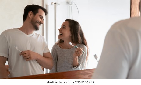 Laughing couple cleaning teeth, holding toothbrushes, standing near mirror in modern bathroom, happy wife and husband enjoying morning routine together, healthcare and oral hygiene concept - Powered by Shutterstock