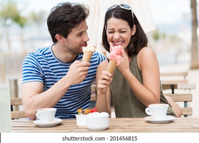 Laughing Couple Eating Ice Cream Cones As They Sit At An Open-air Restaurant Over A Cup Of Coffee In The Summer Sunshine