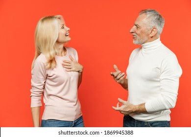 Laughing Couple Friends Elderly Gray-haired Man Blonde Woman In White Pink Casual Clothes Standing Looking At Each Other Speaking Talking Isolated On Bright Orange Color Background Studio Portrait