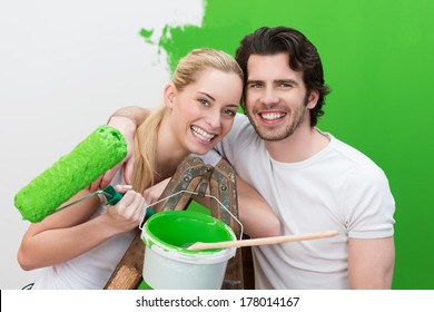 Laughing Couple Painting Their House Green Posing Together In Front Of A Half Finished Wall With A Roller And Tub Of Paint