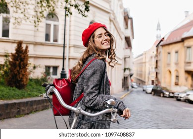 Laughing Curly Dark-haired Girl Looking Back, Standing On Old Street In Morning. Pretty Young Lady With Leather Backpack Posing With Bicycle, Waiting Friend On Avenue.