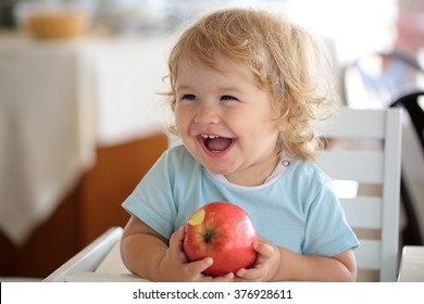 Laughing Cute Fair-haired Blond Hazel-eyed Kid Little Child Baby Boy Sitting In Highchair And Eating Big Red Apple Fruit Portrait On Blurred Background, Horizontal Picture