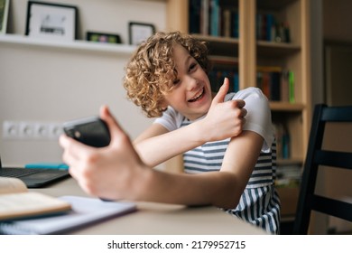 Laughing Cute Little Girl Holding And Using Mobile Phone Showing Thumb Up, Browsing Social Network Apps, Surfing Internet And Scrolling News Feed, Reading Electronic E-book At Table With Laptop.