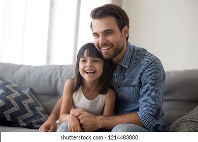 Laughing Diverse Family Sitting On Couch In Living Room At Home. Young Father Little Daughter Having Video Call Using Laptop Or Watching Movie Comedy Or Cartoon. Weekend Activities Free Time Concept