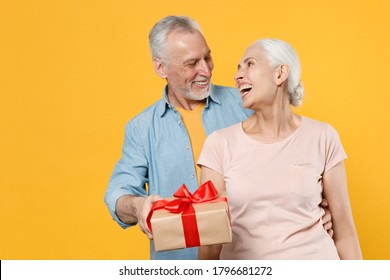 Laughing Elderly Gray-haired Couple Woman Man In Casual Clothes Isolated On Yellow Background Studio. St. Valentine's Day Women's Day, Birthday, Holiday Concept. Hold Present Box With Gift Ribbon Bow