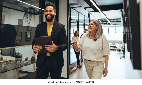 Laughing Entrepreneurs Walking Together In An Office. Two Happy Business Colleagues Sharing A Laugh While Having A Discussion. Diverse Businesspeople Working Together On A Project.