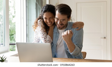 Laughing excited millennial family couple embracing by computer screen winning prize at lottery or sports betting. Loving young wife sharing amazed husband joy of getting reward perfect job proposal