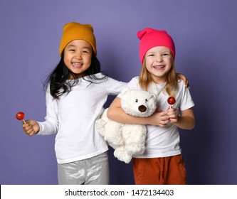 Laughing Friends Kid Girls In White T-shirt And Colorful Pants And Hats Hold Thier Best Friend Polar Bear Toy And Eat Lollipops On Purple. International Day Of Polar Bear Concept.