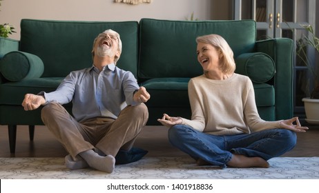 Laughing funny elderly spouses sitting in living room on floor in lotus position practice meditation distracted from yoga exercise joking feels overjoyed, healthy active lifestyle of retirees concept - Powered by Shutterstock