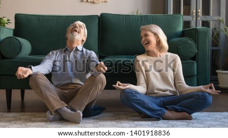 Laughing funny elderly spouses sitting in living room on floor in lotus position practice meditation distracted from yoga exercise joking feels overjoyed, healthy active lifestyle of retirees concept
