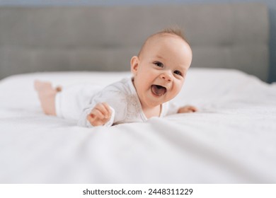 Laughing funny little baby having fun while lying on bed at home. Portrait of happy infant girl in bodysuit resting in bedroom on comfortable blanket. Purity, innocence concept. - Powered by Shutterstock