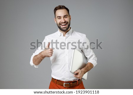 Laughing funny young bearded business man in classic white shirt standing hold laptop pc computer showing thumb up isolated on grey color background studio portrait. Achievement career wealth concept