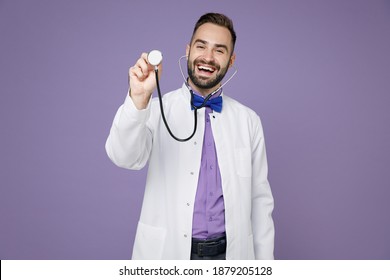 Laughing funny young bearded doctor man wearing white medical gown standing hold stethoscope isolated on violet colour wall background studio portrait. Healthcare personnel health medicine concept