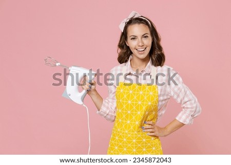 Laughing funny young brunette woman housewife 20s wearing yellow apron hold mixer looking camera while doing housework isolated on pastel pink colour background studio portrait. Housekeeping concept