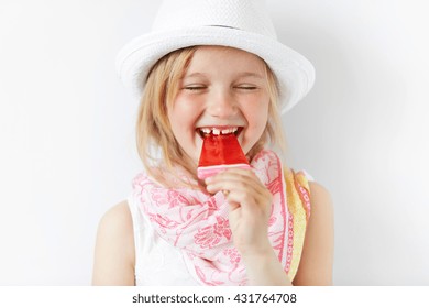 Laughing Girl With Red Popsicle In White Light Room. Cheerful Child Eating Her Sugar Candy With Closed Eyes. Delighted Kid Radiates Positive Emotions And Summer Holiday Mood.