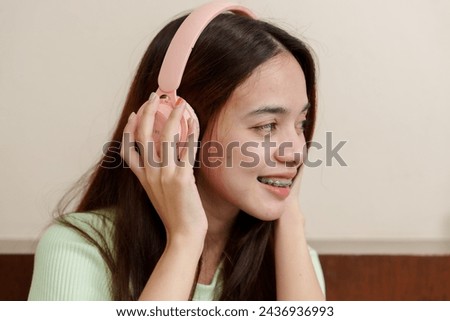Laughing gleefully, Asian female with gleaming braces, rosy headphones in hand, exudes infectious cheerfulness. Radiant with merriment, woman holds pastel headset, teeth adorned with braces