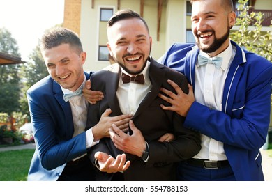 Laughing groomsmen in suits hold smiling groom under his arms