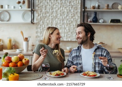 Laughing Happy European Millennial Blond Wife And Hasband With Stubble Eating Pasta In Light Kitchen Interior. Romantic Dinner At Home Together, Love, Relationship And Family During Covid-19 Outbreak