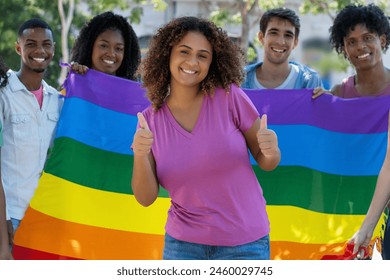 Laughing hispanic female young adult celebrating pride parade with LGBTQ rainbow flag and group of queer people outdoor in summer in city - Powered by Shutterstock