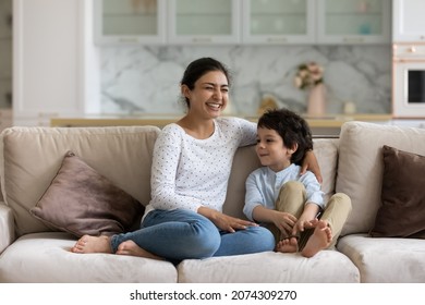 Laughing Indian Mom Enjoying Leisure Time With Happy Son At Home, Talking To Boy. Mother And Cheerful Preschool Child Having Fun Together, Relaxing On Couch. Babysitter Watching Cute Kid