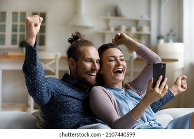 Laughing joyful bonding young family couple looking at smartphone screen, celebrating online lottery giveaway gambling game win, getting message with amazing news or big discount shopping promo code.