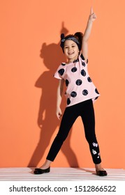 Laughing Kid Girl In Is Standing Holding Her Hand Up, Pointing Up With Her Finger, Showing One Sign, First, Funny Dancing On Orange Background