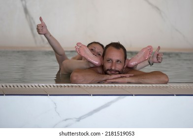 Laughing And Leisure, Couple Enjoying Relaxing In The Swimming Pool. Focus Is On Handsome Muscular Man. Female Feet Above Water. Bearded Man With Has Happy Face. Her Sexy Legs On The Man's Head