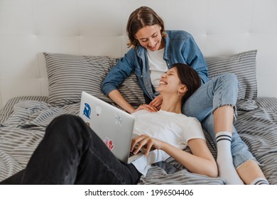 A laughing lesbian couple with laptop looking to each other while lying on the bed in the light room