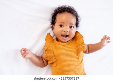 a laughing little African-American baby girl in an orange bodysuit on the bed at home, a funny six-month-old black newborn baby lies on the back and rejoices. View from above. - Powered by Shutterstock