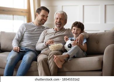 Laughing Little Boy With Grandfather And Father Watching Tv, Eating Popcorn Snack, Sitting On Couch With Soccer Ball, Happy Family Watching Football Game, Spending Leisure Time At Home Together