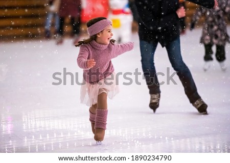 Laughing little girl in a pink sweater is skating on a winter evening on an outdoor ice rink, it is snowing