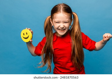 A laughing little girl in red t-shirt is holding a happy emoji emotion icon in her hand and showing thumbs up on blue background. International World smile Day. World emoji day. Birthday Party. - Powered by Shutterstock