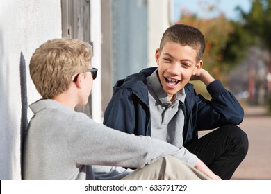 Laughing Male Teenager With Braces Beside Friend Sitting Against Wall Outside
