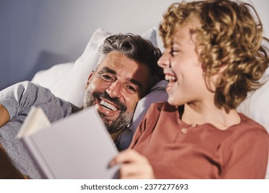 Laughing man and boy bonding while reading interesting book together in bedroom at home